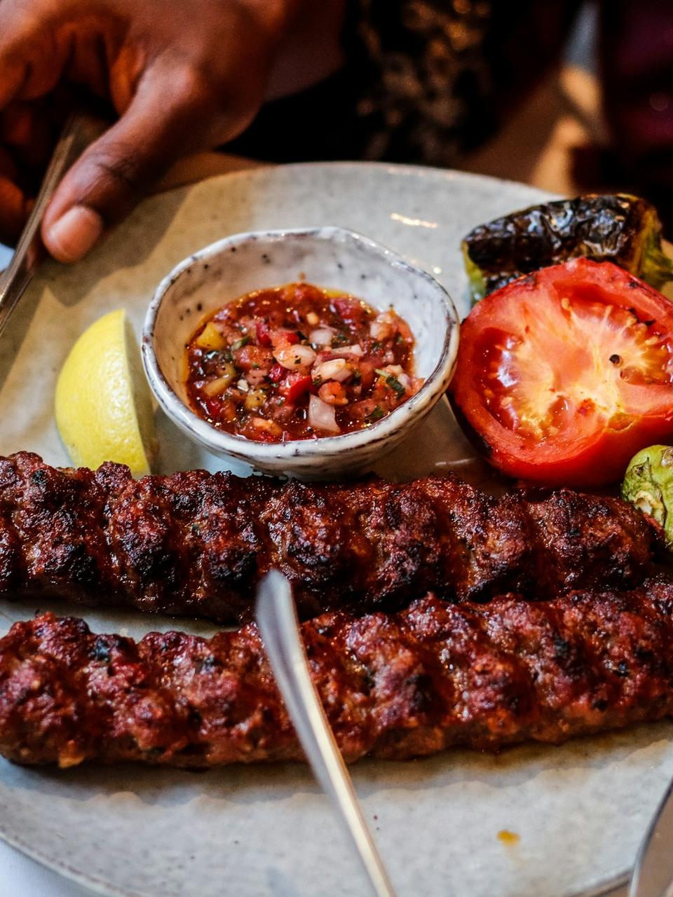 Demo-Foto: Kebabspieße auf einem Teller mit Beilagen, Hände am Tisch