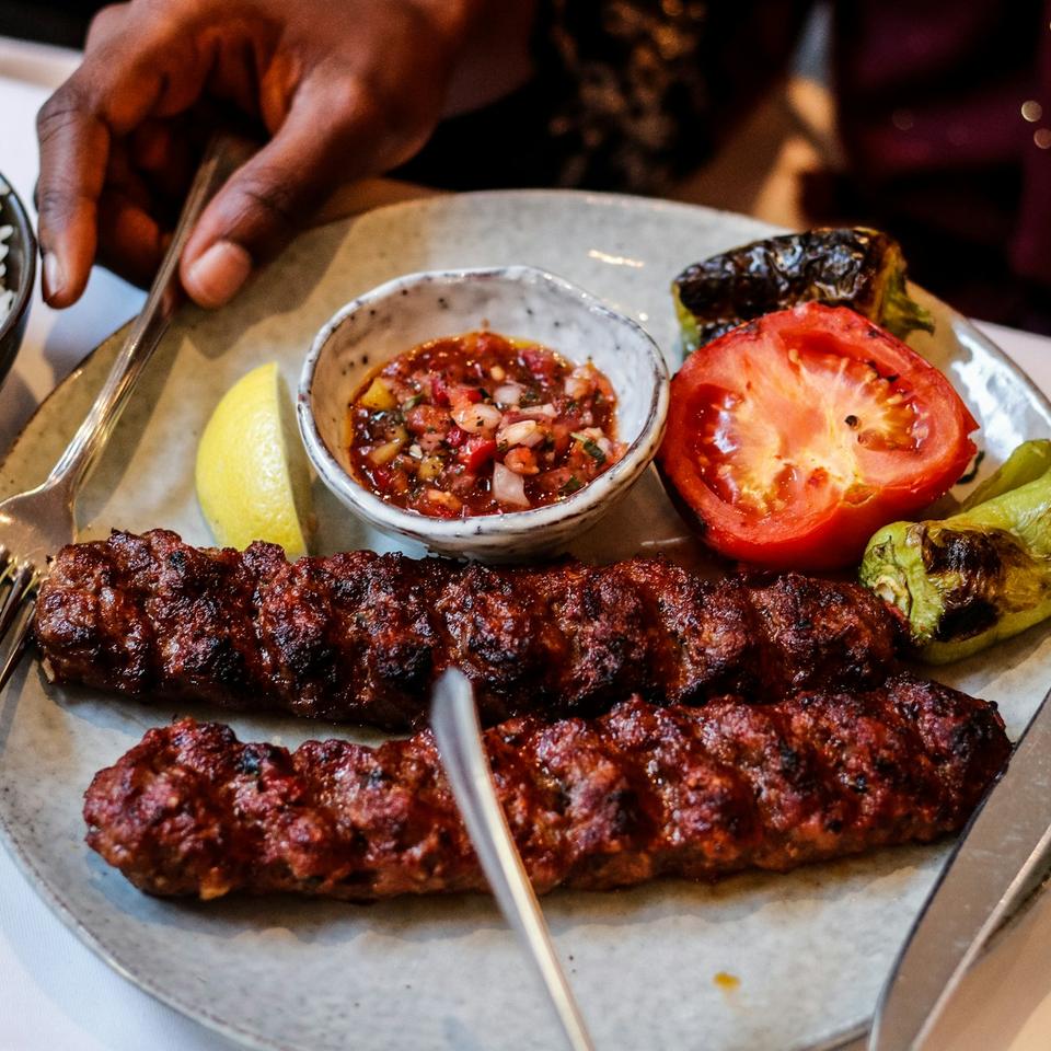 Demo-Foto: Kebabspieße auf einem Teller mit Beilagen, Hände am Tisch