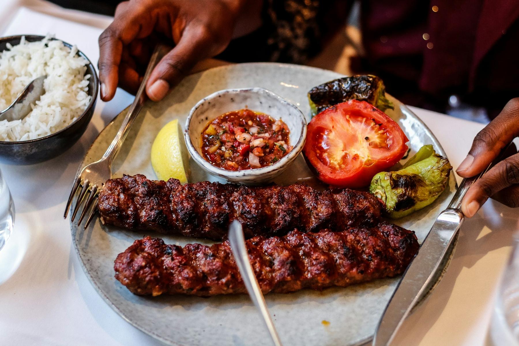 Demo-Foto: Kebabspieße auf einem Teller mit Beilagen, Hände am Tisch